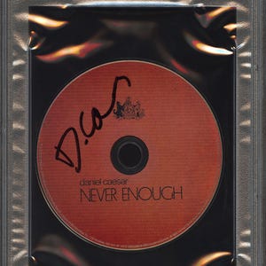 May include: A Daniel Caesar "Never Enough" CD, encased in a protective plastic holder. The CD is orange with black text and a signature. The holder has a PSA/DNA certification label.