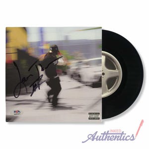 May include: A signed vinyl record with a black disc and a cover featuring a blurred image of a person running. The cover includes the text "Parental Advisory Explicit Content" and a signature. The record is from Inked Authentics.