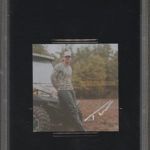 May include: A PSA/DNA certified CD insert featuring Tucker Wetmore. The insert shows a man in a camouflage shirt and a baseball cap leaning against a vehicle. The background includes trees and a field.