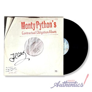 May include: A vintage vinyl record album by Monty Python, titled "Contractual Obligation Album." The album cover is off-white with red text and a signature. A black vinyl record is visible on the right side of the image. The album is from RCA.