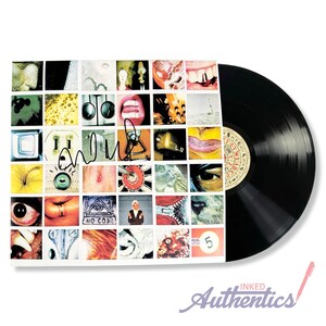 May include: A black vinyl record next to a record sleeve with a grid of colorful, abstract images. The sleeve is signed with a black marker. The record label is visible. The words "INKED Authentics" are at the bottom.