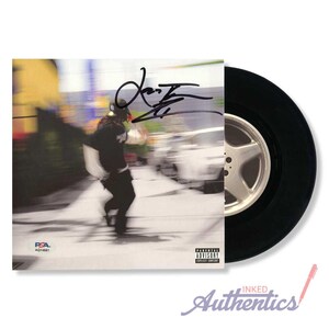 May include: A black vinyl record with a white and silver wheel design. The album cover features a blurred image of a person running, with a signature and the text "Parental Advisory Explicit Content".
