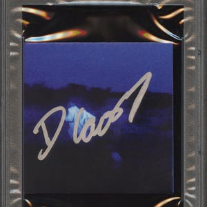 May include: A CD cover featuring a dark blue image with a signature in silver ink. The CD cover is encased in a clear plastic holder with a label at the top that reads "CD COVER DANIEL CAESAR PSA/DNA CERTIFIED AUTHENTIC AUTO".