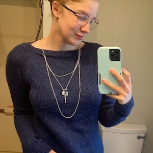 Könnte beinhalten: Ein marineblauer Strickpullover mit Rundhalsausschnitt und langen Ärmeln. Der Pullover wird mit einer silbernen Halskette mit Anhänger kombiniert. Die Person trägt eine Brille und hält ein hellgrünes Telefon.