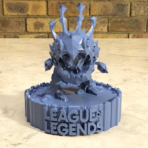 May include: A light blue, 3D-printed figure of a creature with a spiky crown and jagged features. The figure is standing on a circular base with the words "LEAGUE OF LEGENDS" in raised letters. The figure is set against a neutral background.