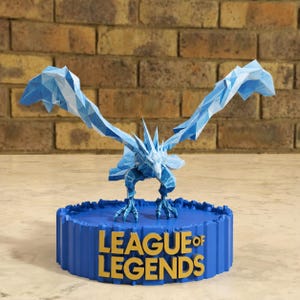 May include: A low-poly papercraft figure of a blue ice dragon with outstretched wings, mounted on a circular blue base. The base features the gold-colored text "LEAGUE OF LEGENDS" in a bold, sans-serif font, displayed prominently on the front.