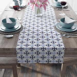 May include: A white table runner with a geometric pattern of blue and black crosses. The runner is laid out on a wooden table with place settings and a vase of pink flowers.
