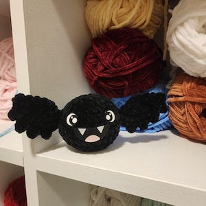 Peut inclure: Une chauve-souris en crochet noir avec un visage souriant et des yeux blancs. La chauve-souris est posée sur une étagère blanche avec d'autres pelotes de laine en arrière-plan.