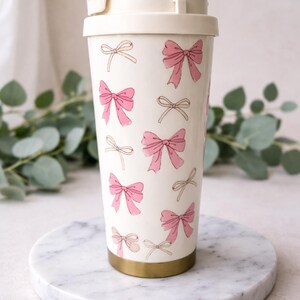 Könnte beinhalten: Ein weißer Reisebecher mit cremefarbenem Deckel und goldfarbenem Boden. Der Becher ist mit rosa und cremefarbenen Schleifenillustrationen verziert. Der Becher steht auf einer weißen Marmoroberfläche.