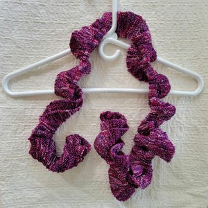 May include: A knitted infinity scarf in shades of purple, pink, and white hangs on a white plastic hanger. The scarf has a unique, ruffled design. The yarn appears to be a soft, textured material, perfect for cold weather.