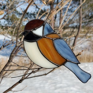 May include: A stained glass bird ornament in shades of brown, white, black, and blue. The bird is perched on a thin metal wire and a small branch. The background shows a snowy landscape and bare tree branches.