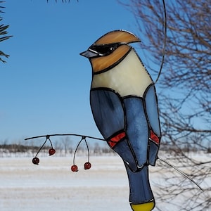 Peut inclure: Sculpture d'oiseau en vitrail, perché sur une branche métallique avec des baies rouges. L'oiseau a une poitrine crème, des ailes bleues et une queue jaune. La tête est beige et noire, sur fond de ciel bleu clair.
