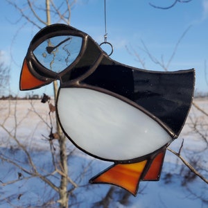 May include: Stained glass puffin bird suncatcher with black, white, orange, and yellow details. The bird is suspended by a thin wire against a blue sky and winter landscape. The bird's eye reflects the sky.