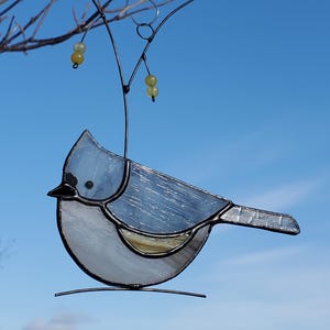 May include: A stained glass bird suncatcher with a light blue body, black beak, and a wire hanger. The bird is perched on a wire and has a yellow bead accent. The background is a clear blue sky.