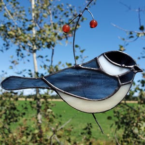 May include: A stained glass bird suncatcher with a blue and white body, black head, and a tail. The bird is perched on a thin metal wire with two red glass berries. The background features a green field and a blue sky.