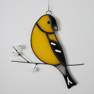 May include: Stained glass bird decoration. A yellow bird with black and white details perches on a thin black branch with white berries. The bird has a black head and tail, and is suspended from a black wire.