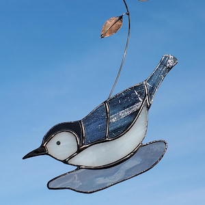 May include: A stained glass bird sculpture with a white breast, blue back, and black head. The bird is perched on a clear glass base and is suspended from a thin wire with two copper-coloured leaves. The background is a clear blue sky.