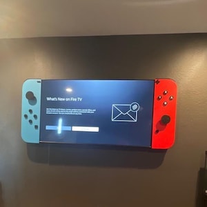 May include: A large television mounted on a dark wall, designed to resemble a video game controller. The screen displays "What's New on Fire TV." A colorful star-patterned rug is on the floor. A ladder and aquarium are also visible.