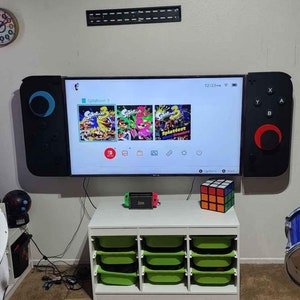 May include: A large television screen modified to resemble a Nintendo Switch controller, mounted on a wall. The screen displays the game "Splatoon 3." Below, a white storage unit with green bins holds a Nintendo Switch console and a Rubik's Cube. A drum set is visible.