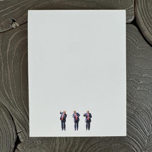 May include: A white greeting card with three small figures in suits and red ties. The figures are in different poses. The card is on a textured, dark gray surface.