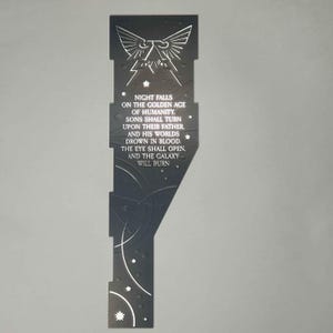 Könnte beinhalten: Ein schwarzes Metall-Lesezeichen mit einem weißen Adler-Design und einem Zitat über den Fall der Menschheit. Das Zitat lautet: "Die Nacht bricht herein über das goldene Zeitalter der Menschheit. Söhne werden sich gegen ihren Vater wenden, und seine Welten ertrinken in Blut. Das Auge wird sich öffnen, und die Galaxie wird brennen."