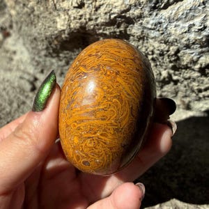 May include: A polished, oval-shaped stone with a swirling pattern of orange and brown hues. The stone's surface is smooth and reflective, showcasing the natural variations in color and texture. The stone is held in a hand.