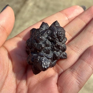May include: A dark, textured mineral specimen with a rough, crystalline surface. The mineral is a deep, charcoal gray color and appears to be held in a hand. The overall shape is irregular and clustered, with multiple points and facets.