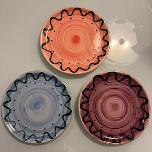May include: Three ceramic plates with a wavy edge and black and green painted designs. The plates are in shades of pink, blue, and purple.