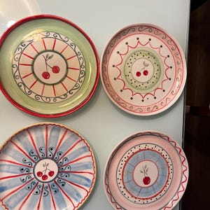 Puede incluir: Cuatro platos decorativos de cerámica con diseños de cerezas. Cada plato presenta una combinación de colores diferente, incluyendo verde, rojo, azul y rosa, con un motivo central de cereza y bordes estampados. Los platos están dispuestos sobre una superficie reflectante.