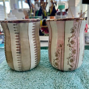 Puede incluir: Dos tazas de cerámica con ribetes dorados y patrones intrincados. Una taza tiene un patrón de rayas rosa y blanco con acentos florales dorados. La otra taza tiene un patrón de rayas marrón y blanco con acentos dorados.