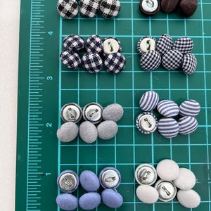May include: An assortment of fabric-covered buttons in different colours and patterns, such as black and white gingham, navy and white checks, and solid grey, blue, and white. The buttons are arranged on a green cutting mat with inch and cm measurements.