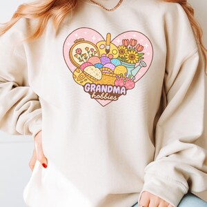 May include: A beige sweatshirt featuring a heart-shaped graphic with the words "Grandma hobbies." The graphic includes a basket of yarn, embroidery hoop, paint palette, flowers, and a watering can. The sweatshirt has long sleeves.