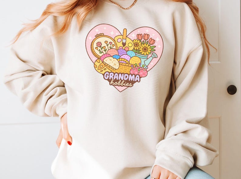 May include: A beige sweatshirt featuring a heart-shaped graphic with the words "Grandma hobbies." The graphic includes a basket of yarn, embroidery hoop, paint palette, flowers, and a watering can. The sweatshirt has long sleeves.
