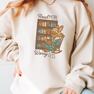 May include: Beige sweatshirt featuring a cartoon rabbit reading a book in a rocking chair, with the text "Read More Worry Less." The rabbit wears glasses and a bonnet, holding a cup. A bookshelf is behind the rabbit.