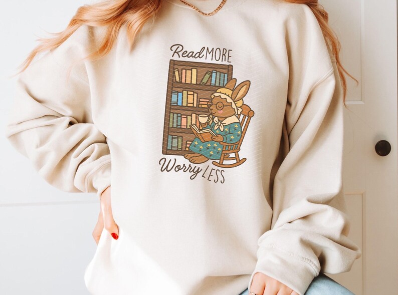 May include: Beige sweatshirt featuring a cartoon rabbit reading a book in a rocking chair, with the text "Read More Worry Less." The rabbit wears glasses and a bonnet, holding a cup. A bookshelf is behind the rabbit.