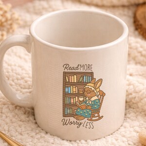 May include: Cream-colored ceramic mug with a whimsical illustration of a bunny reading in a rocking chair, next to a bookshelf. The mug has the text "Read More Worry Less". The mug has a curved handle.