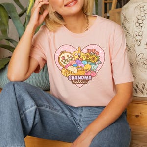May include: A light pink t-shirt featuring a heart-shaped graphic with the words "Grandma hobbies." The graphic includes images of embroidery, paintbrushes, flowers, and yarn. The shirt is a crew neck style.