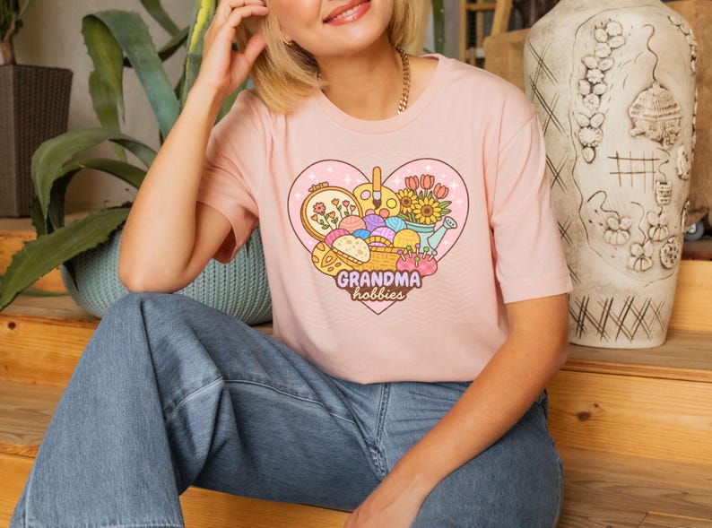 May include: A light pink t-shirt featuring a heart-shaped graphic with the words "Grandma hobbies." The graphic includes images of embroidery, paintbrushes, flowers, and yarn. The shirt is a crew neck style.