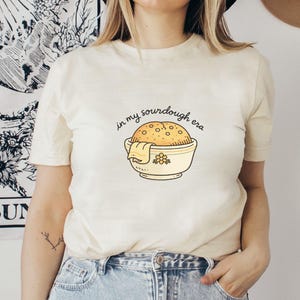 May include: Cream-colored t-shirt with the phrase "in my sourdough era" above a cartoon illustration of a sourdough loaf in a bowl. The bowl is yellow with a floral design. The shirt is short-sleeved.