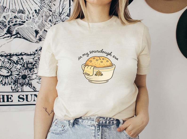 May include: Cream-colored t-shirt with the phrase "in my sourdough era" above a cartoon illustration of a sourdough loaf in a bowl. The bowl is yellow with a floral design. The shirt is short-sleeved.