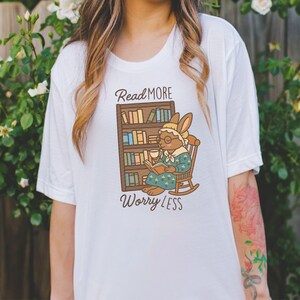 May include: White t-shirt featuring a cartoon rabbit reading a book in a rocking chair, with a bookshelf behind it. The text "Read More Worry Less" is above and below the illustration. The shirt is a casual, comfortable style.