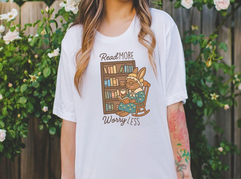 May include: White t-shirt featuring a cartoon rabbit reading a book in a rocking chair, with a bookshelf behind it. The text "Read More Worry Less" is above and below the illustration. The shirt is a casual, comfortable style.
