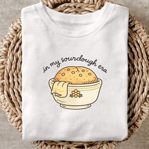 May include: White t-shirt with a sourdough bread graphic in a bowl, with the text "in my sourdough era". The bread is golden brown with a textured surface. The bowl is cream-colored with a floral design.
