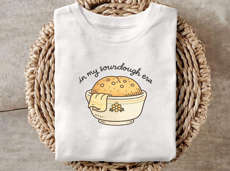 May include: White t-shirt with a sourdough bread graphic in a bowl, with the text "in my sourdough era". The bread is golden brown with a textured surface. The bowl is cream-colored with a floral design.