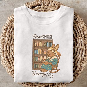 May include: White t-shirt with a graphic of a bunny reading a book in a rocking chair, with a bookshelf behind it. The text "Read More Worry Less" is above and below the image. The shirt is displayed on a woven basket.