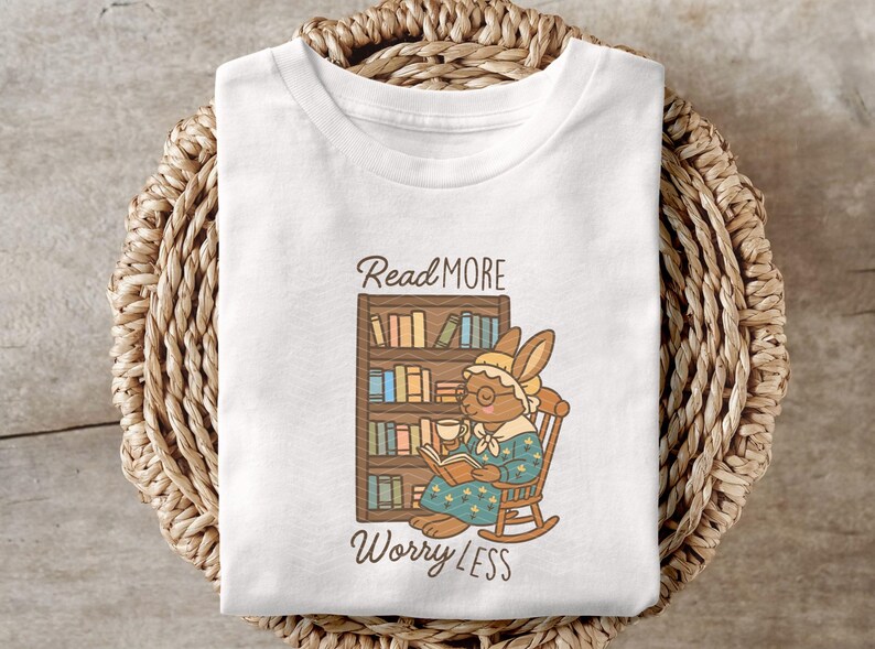 May include: White t-shirt with a graphic of a bunny reading a book in a rocking chair, with a bookshelf behind it. The text "Read More Worry Less" is above and below the image. The shirt is displayed on a woven basket.