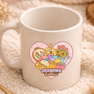 May include: A cream-colored ceramic mug with a heart-shaped graphic featuring craft supplies. The graphic includes embroidery, paintbrushes, yarn, flowers, and the text "grandma hobbies". The mug has a curved handle.