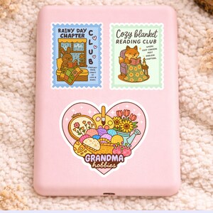 May include: A pink phone case adorned with three stickers. The top stickers read "Rainy Day Chapter Club" and "Cozy Blanket Reading Club." The bottom sticker is heart-shaped, with the text "Grandma hobbies."