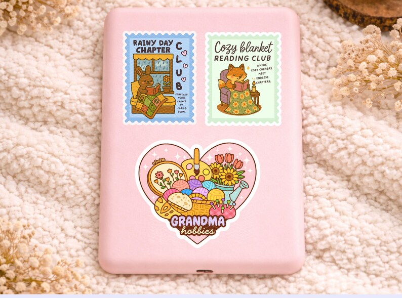 May include: A pink phone case adorned with three stickers. The top stickers read "Rainy Day Chapter Club" and "Cozy Blanket Reading Club." The bottom sticker is heart-shaped, with the text "Grandma hobbies."