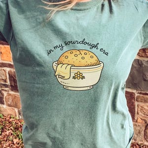 May include: A teal t-shirt with the phrase "in my sourdough era" above a graphic of a sourdough loaf in a bowl. The bowl has a floral design and a cloth draped over the side.